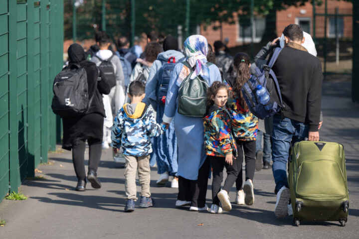 Un grupo de refugiados, incluidos niños y adultos, camina cargando mochilas y maletas. La imagen refleja familias en tránsito hacia un refugio o centro de acogida.