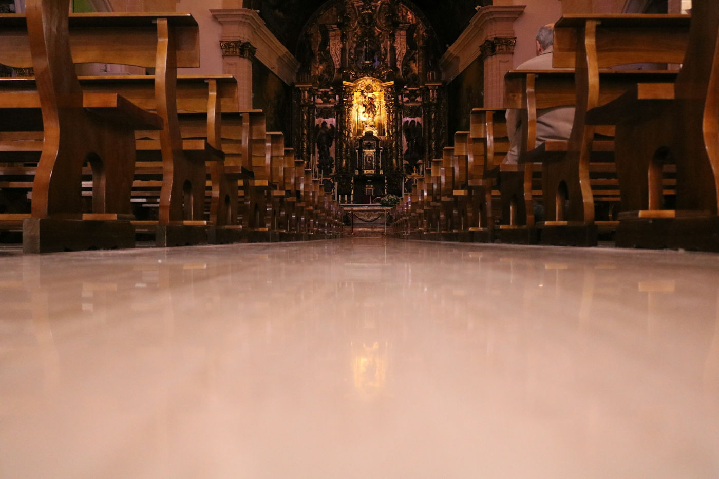 Vista desde el suelo de los bancos de una iglesia vacía, enfocando hacia el altar iluminado al fondo.